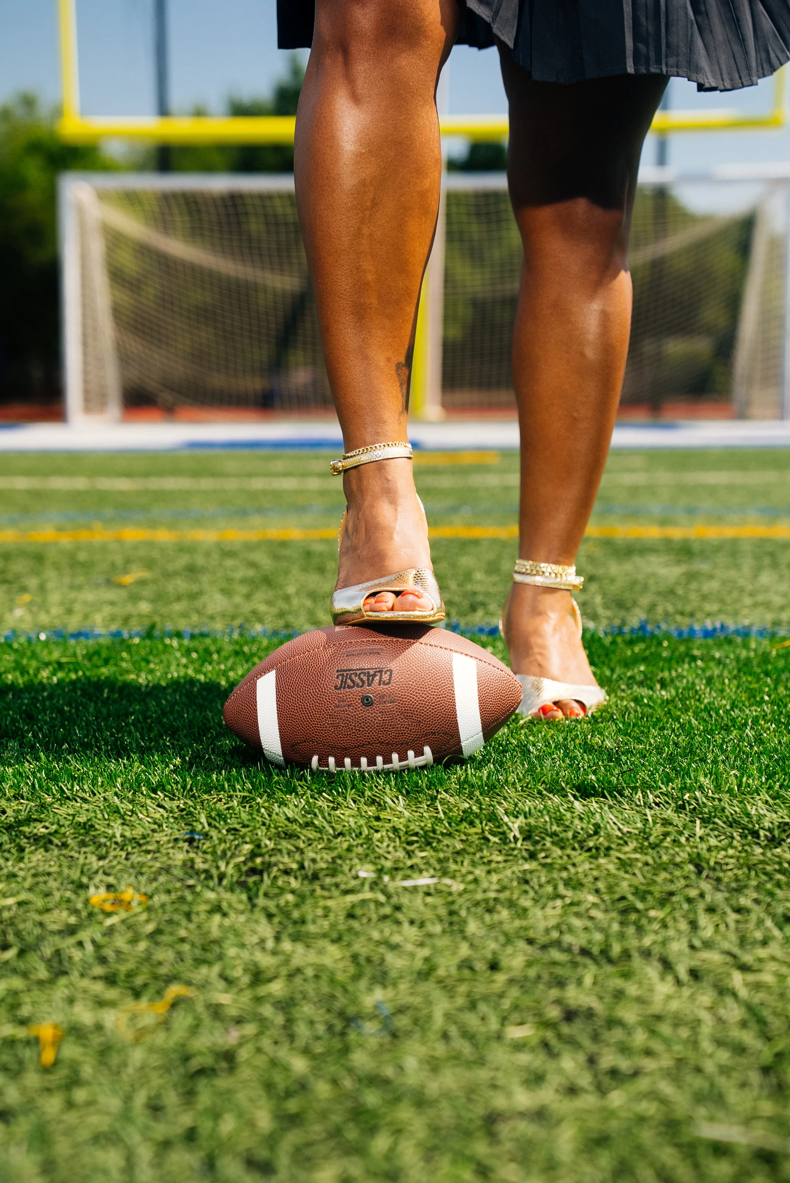 A gold heel resting on a football on a turf field, goalposts out of focus behind.